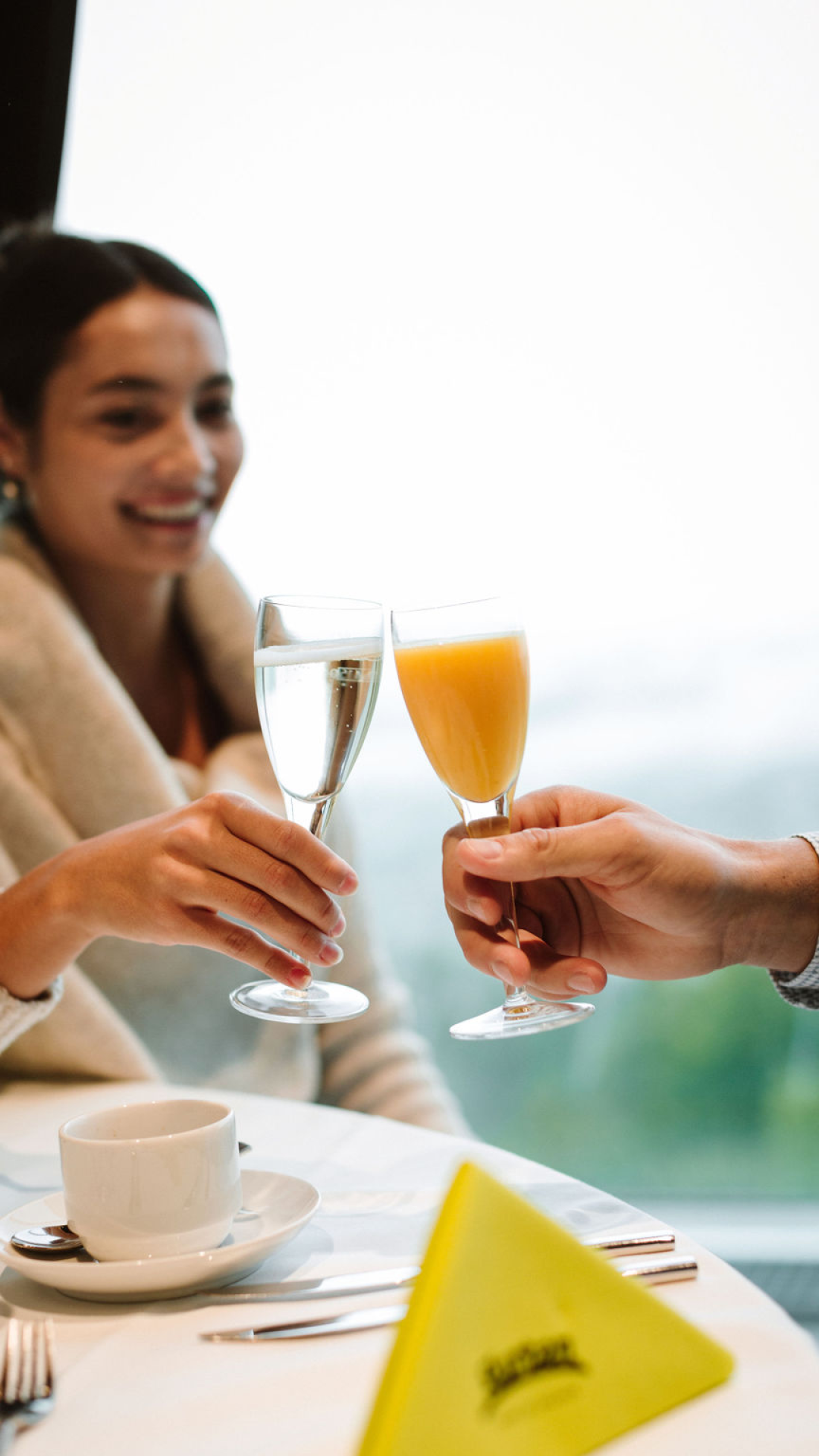 Zwei Personen welche mit einem Champagnerglas anstossen. Die eine Person stosst mit Orangensaft an und die sichtbare Person auf der linken Seite mit Champagner.