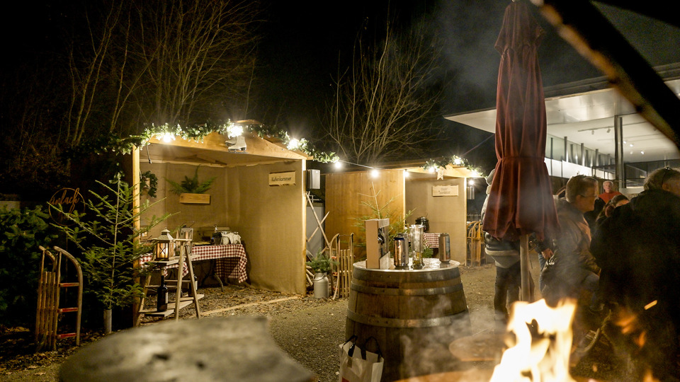 Winterdorf auf dem Gurten mit beleuchteten Holzhütten, Feuerstelle und Gästen bei Nacht.