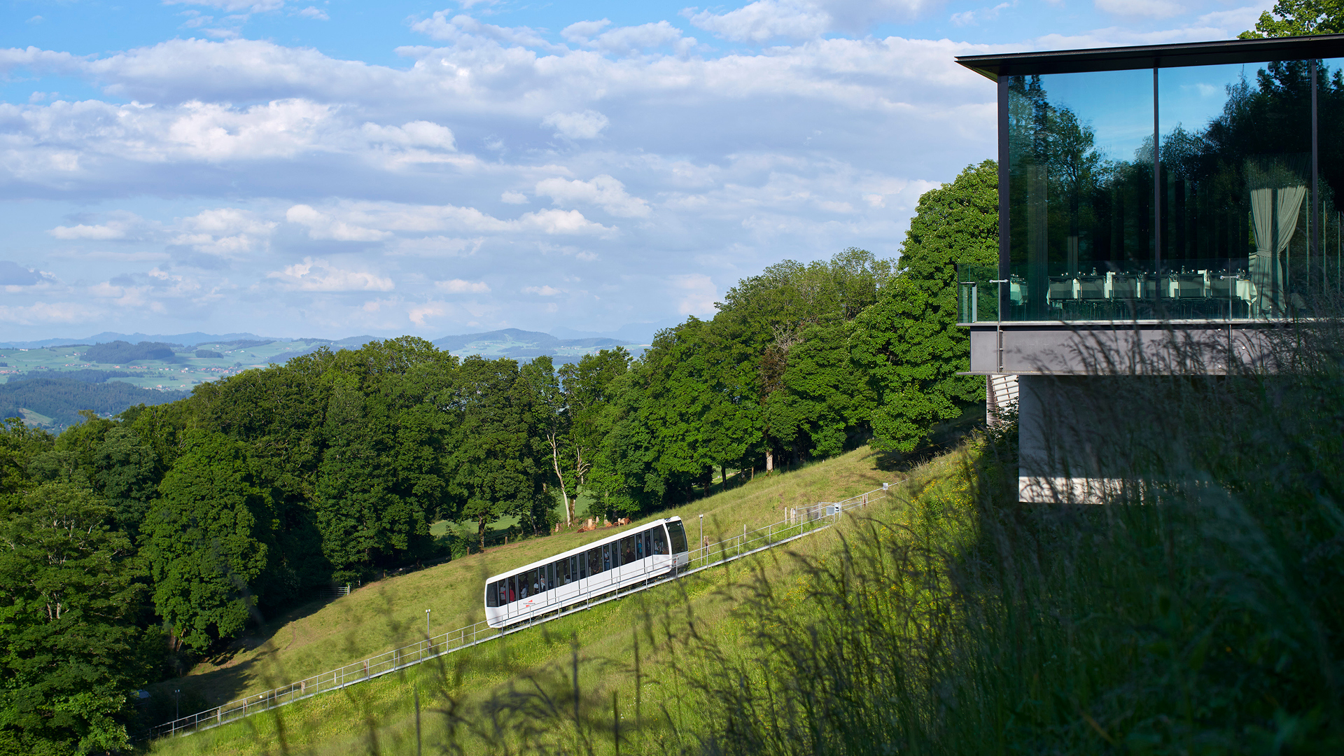 Die Gurtenbahn vor einem Wald und neben dem Eventlokal Pavillon aus dem Gras aufgenommen.