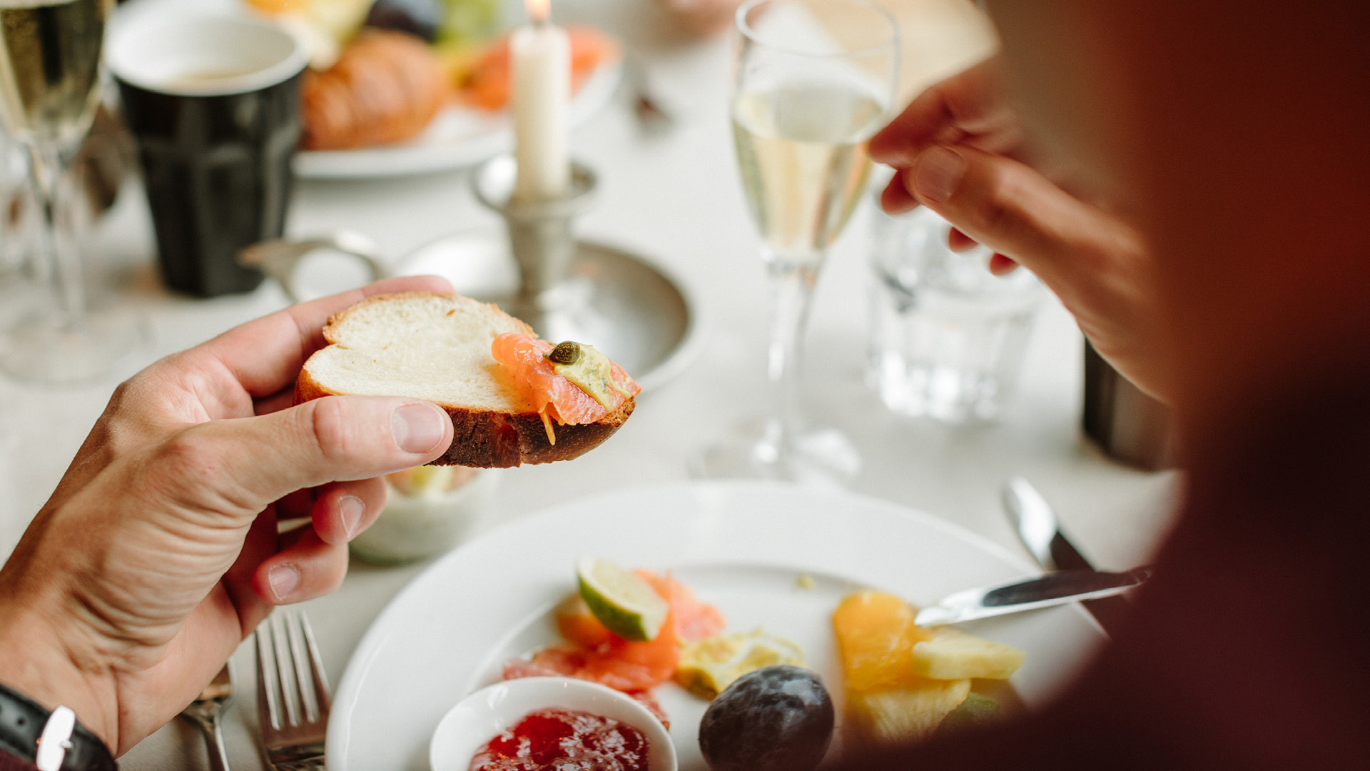 Eine Person, welche ein Stück Brot mit Lachs belegt beim Sunntigsbrunch in der Hand hält. Auf ihrem Teller befindet sich noch mehr Lachs und Früchte.