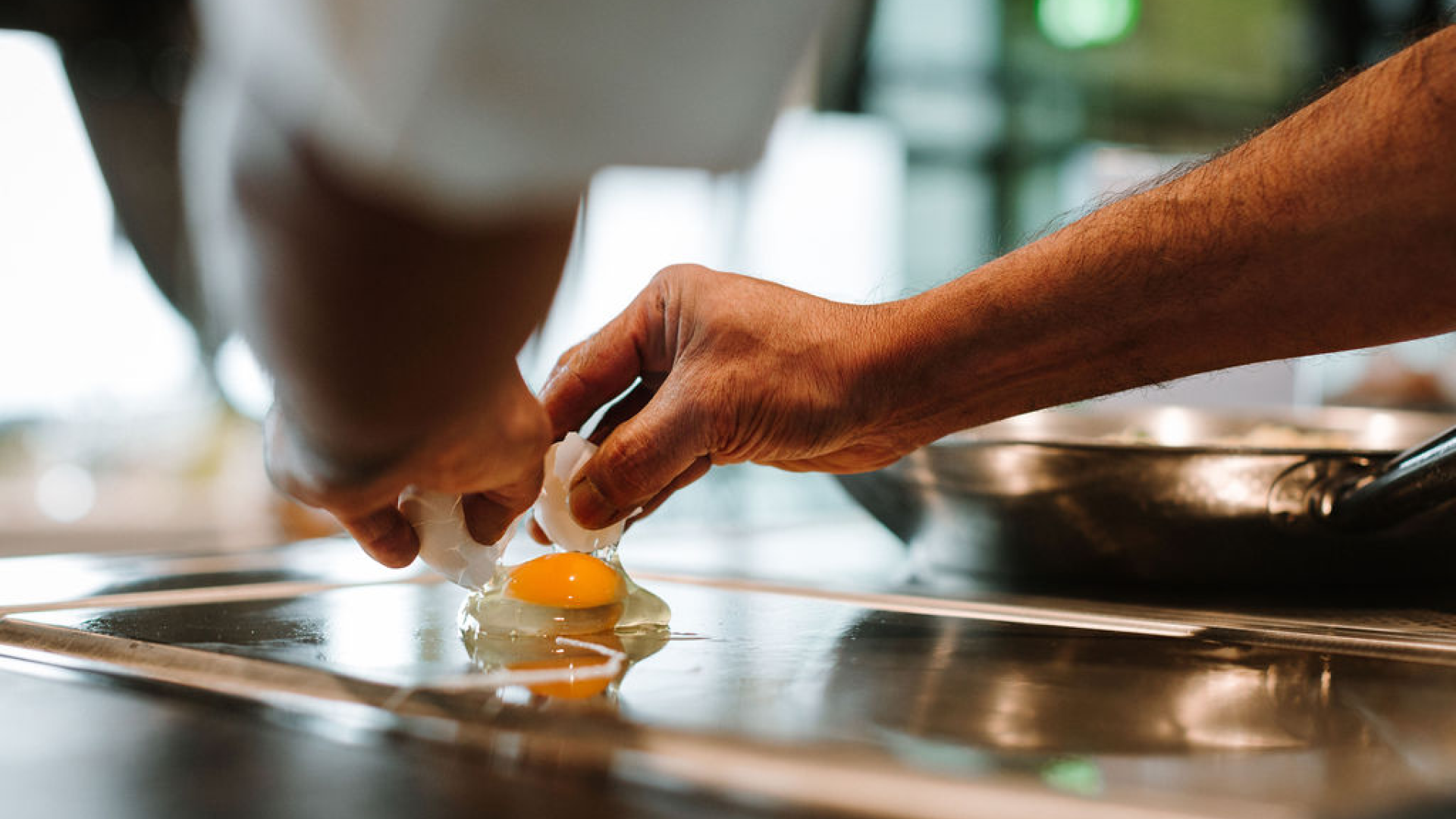 Eine Person welche ein rohes Ei auf einer Pfanne aufschlägt um es zu braten.