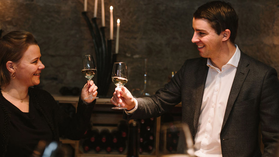 Zwei Personen stossen im Weinkeller mit einem Glas Weisswein an.