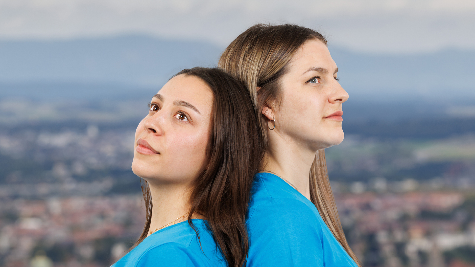 Zwei junge Frauen stehen Rücken an Rücken aneinander. man sieht nur ihre Köpfe. Dahinter die Aussicht vom Gurten auf die Stadt.