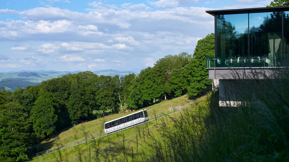 Die Gurtenbahn vor einem Wald und neben dem Eventlokal Pavillon aus dem Gras aufgenommen.