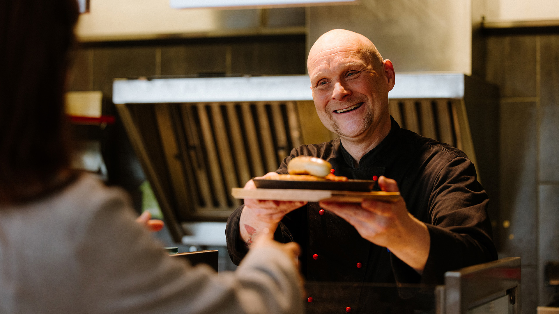Ein Koch mit schwarzer Kochschürze gibt einem Gast eine Rösti