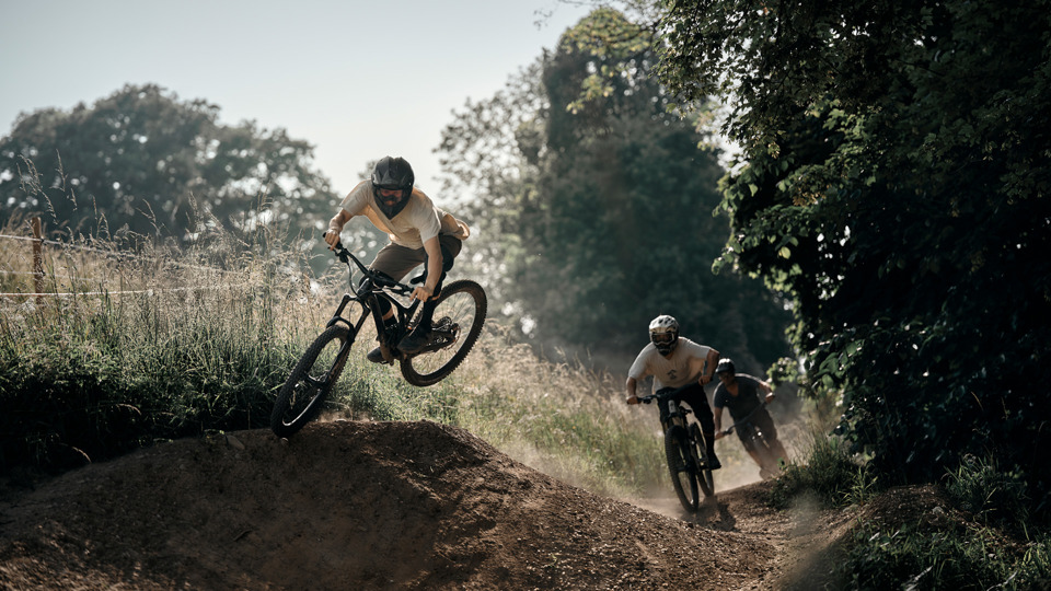 Drei Mountainbiker fahren Gurtentrail herunter, einer springt
