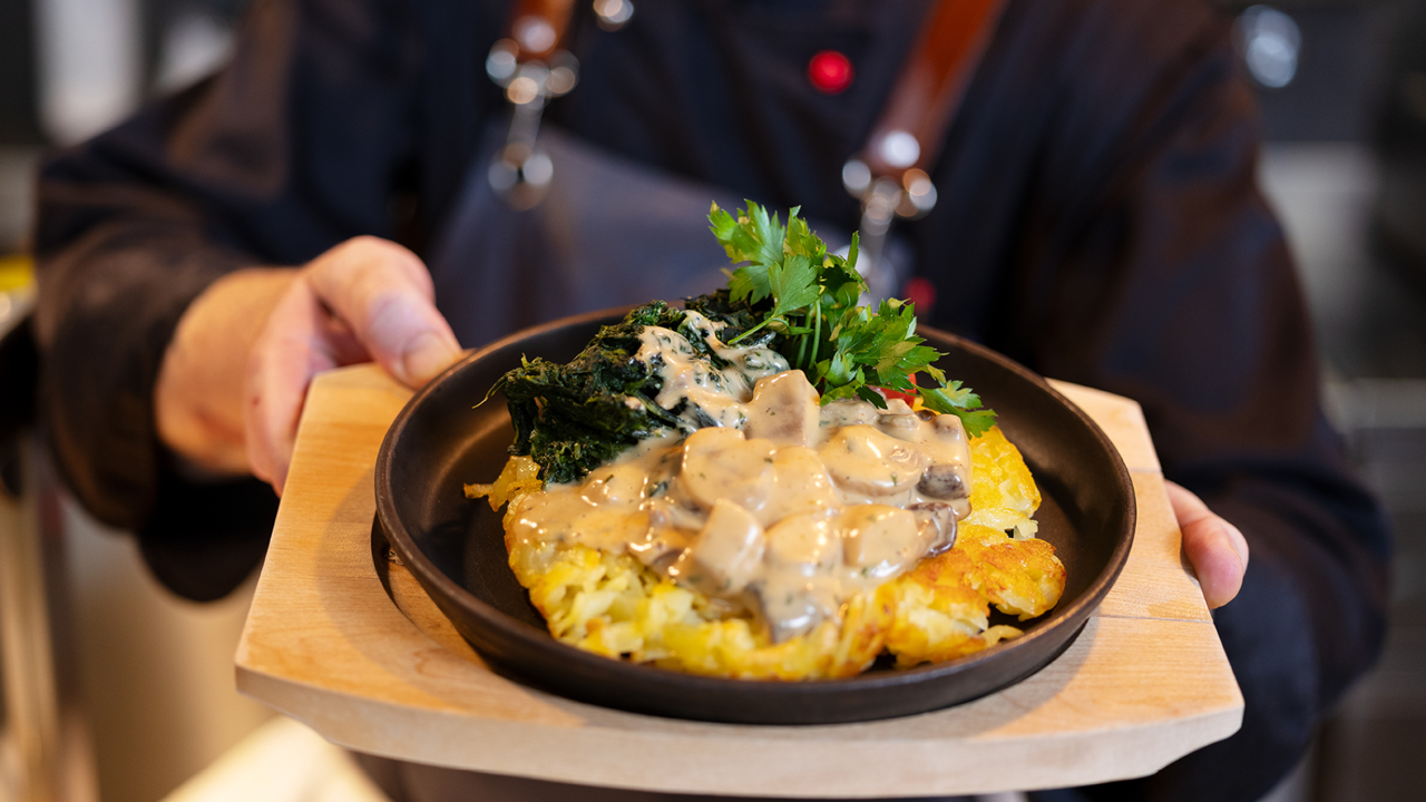 Ein Koch mit einer schwarzen Kochjacke und einer grauen Schürze welcher eine Rösti auf einem schwarzen Teller in der Hand hält. Die Beilage zu Rösti ist Spinat und eine Pilzsauce.