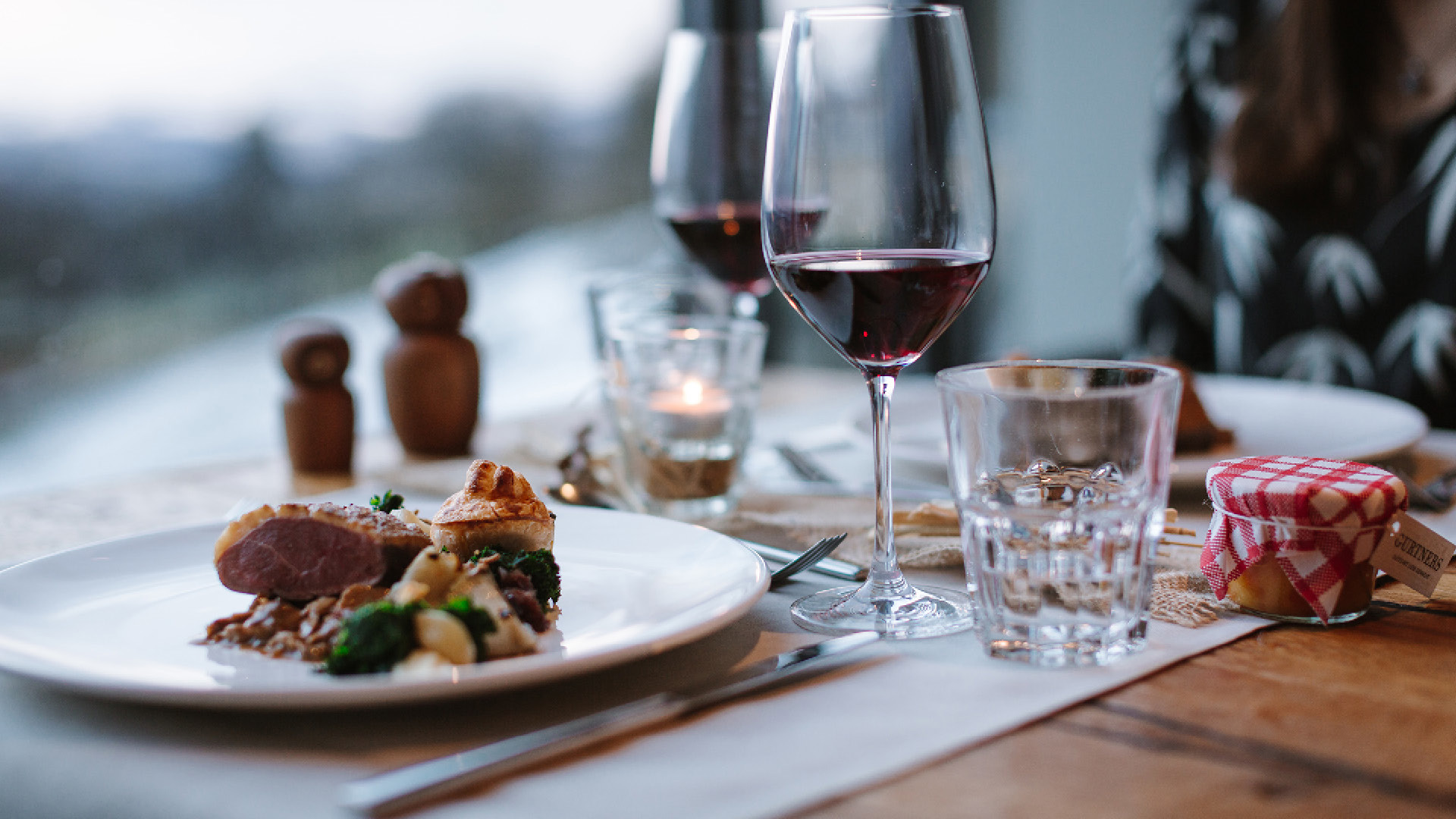 Ein Tisch im Restaurant Gurtners mit zwei Tellern, zwei Weingläsern und einem Willkommensgeschenk. Die Teller sind mit Fleisch und Gemüse bestückt.