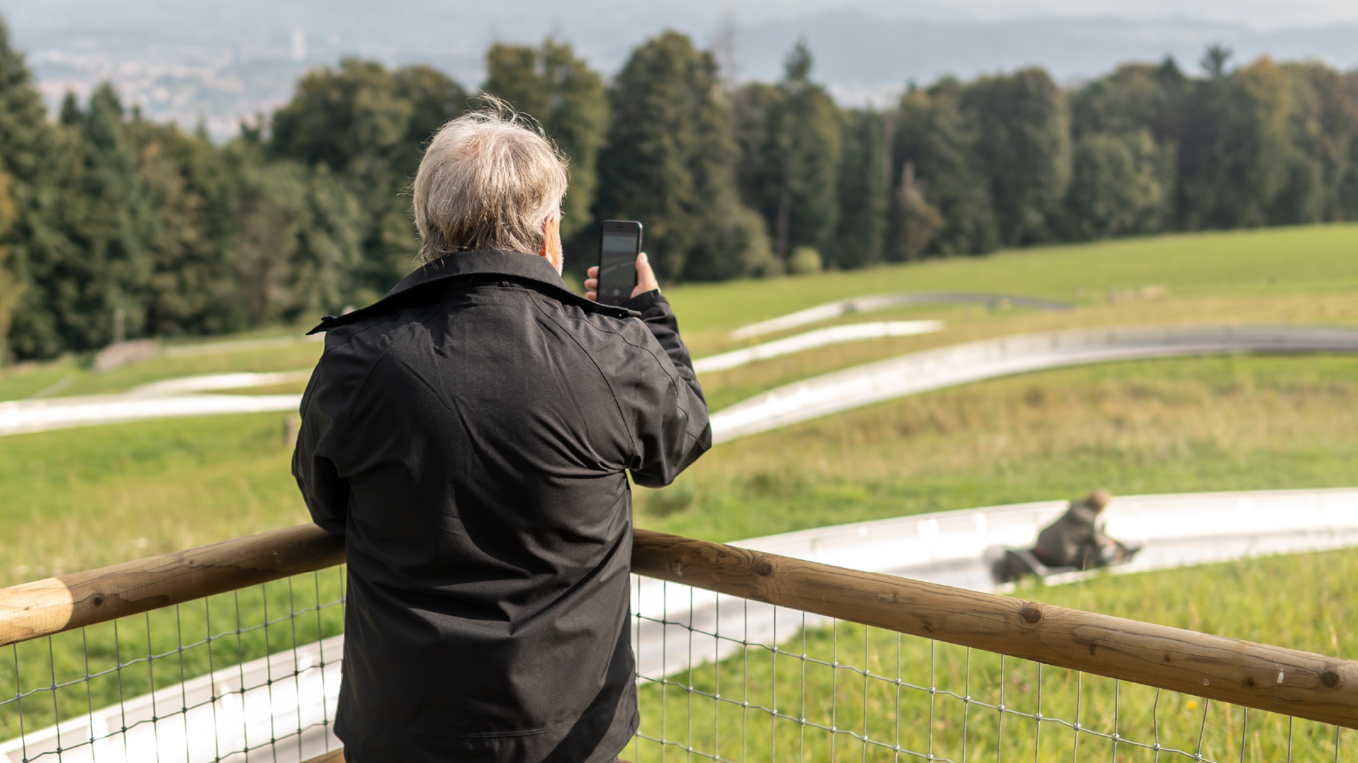 Grossvater von hinten, der am Fotopoint der Rodelbahn seinen Enkel beim rodeln fotografiert