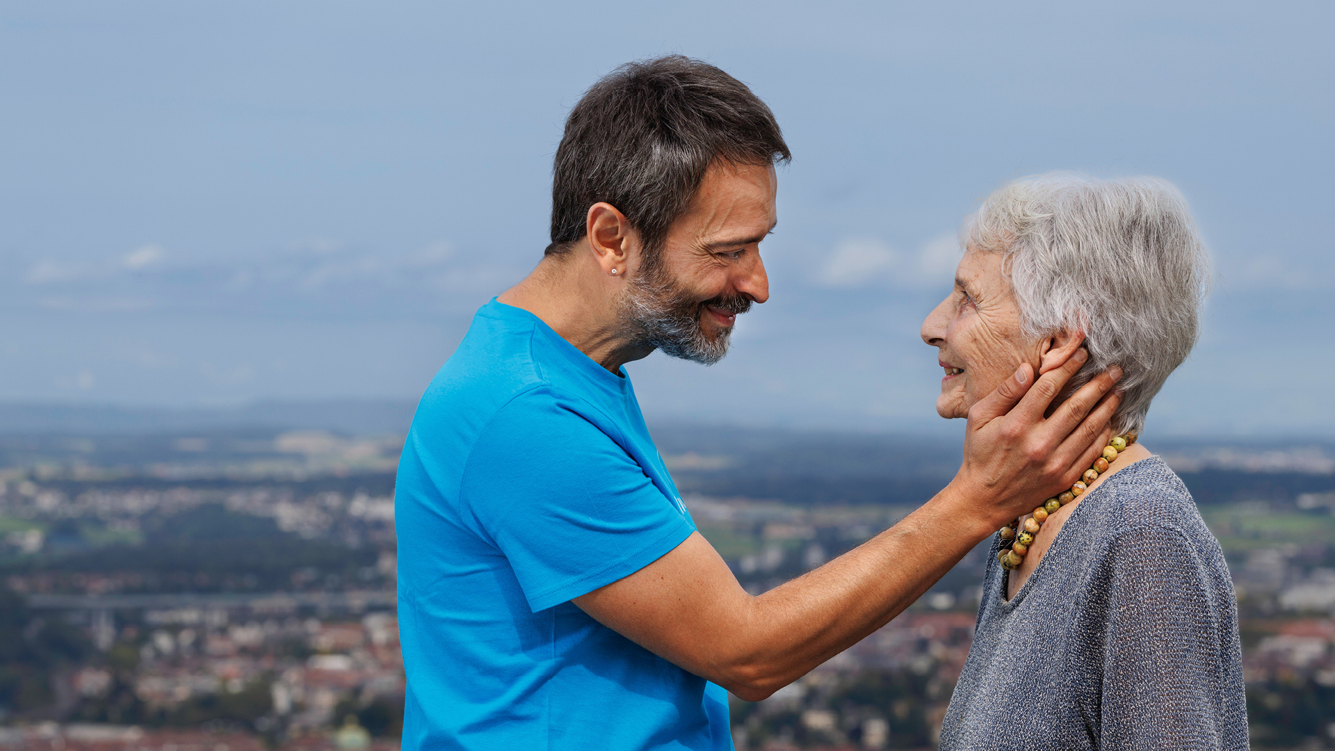 Ein Mann mit Bart legt liebevoll seine Hand auf die Backe einer älteren Frau. Dahinter die Aussicht vom Gurten auf Bern.