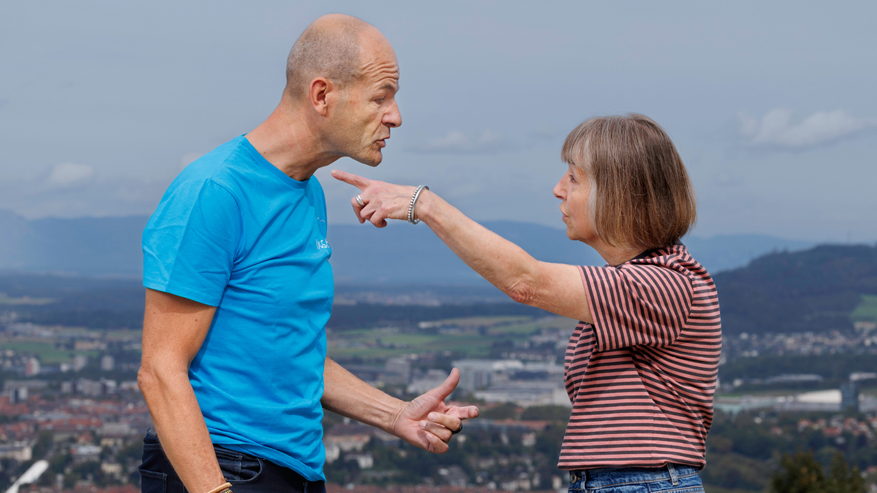 Eine Frau und ein Mann streiten sichtlich. Die Frau macht mit der Hand eine wegweisende Geste. Dahinter die Aussicht auf die Stadt Bern.