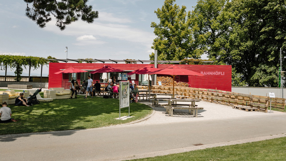 Das Restaurant Bahnhöfli. Es ist rot gestrichen, hat bei den Tischen rote Sonnensschirme und liegt neben einer grünen Wiese.