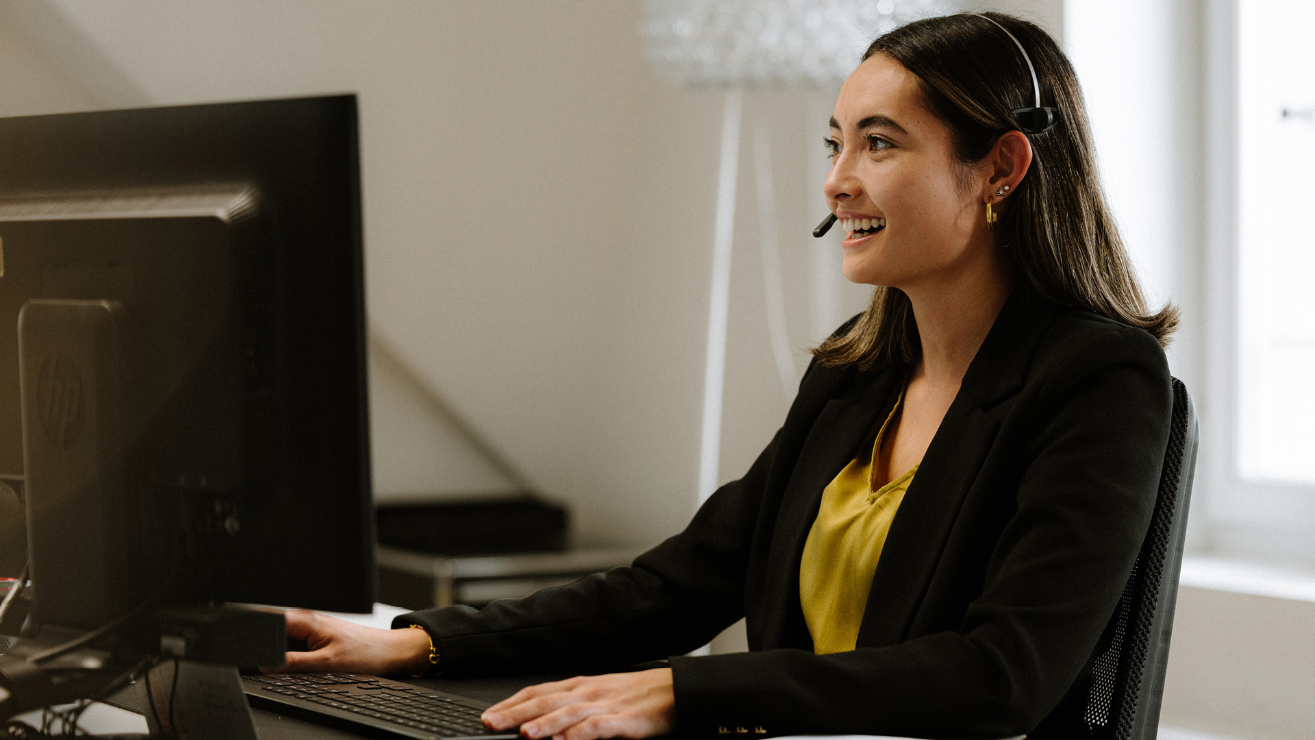 Eine Person in einer gelben Blouse und einem schwarzen Blazer, sitzt an einem Pult mit einem Computer. Sie trägt ein Headset und schreibt etwas auf der Tastatur.