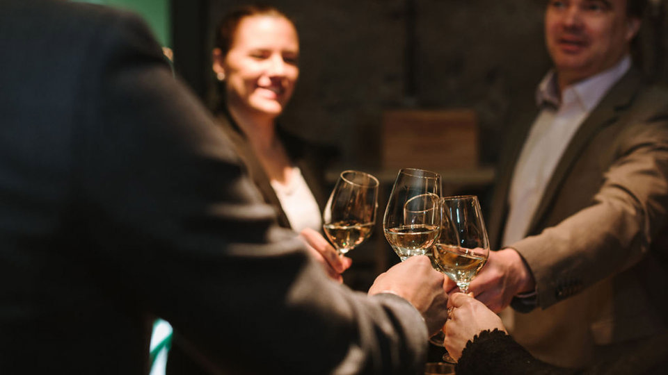 Vier Personen stossen im Weinkeller mit einem Glas Weisswein an.