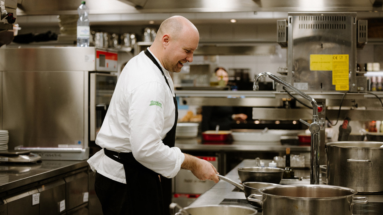 Ein Koch in einer weissen Kochjacke und einer schwarzen Schürze, welcher eine Pfanne schwenkt. Er steht vor dem Kochfeld in der Küche des Gurtners.