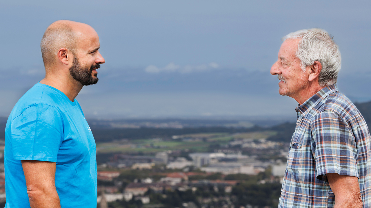 junger Mann und alter Mann stehen sich gegenüber. Dahinter die Aussicht vom Gurten auf Berns Agglomeration.