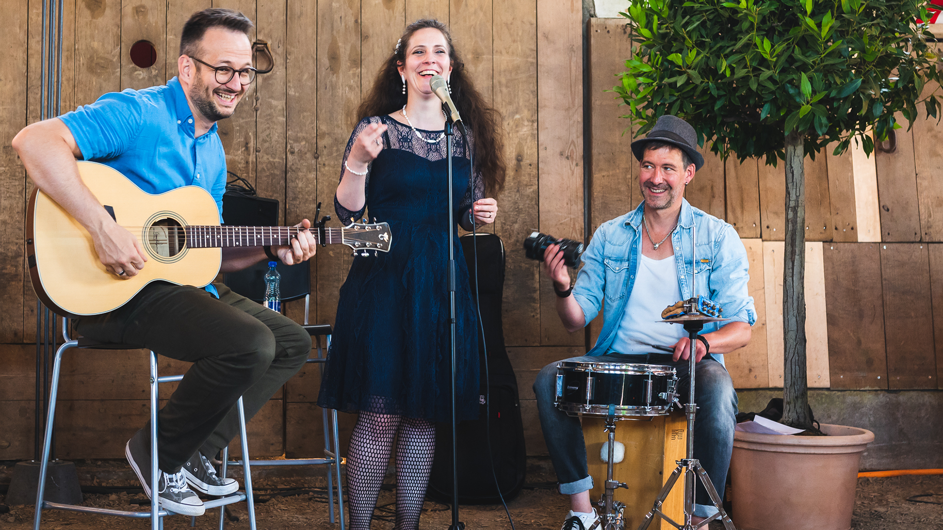 Eine Sängerin, ein Gitarrist und ein Schlagzeuger vor einer Holzwand