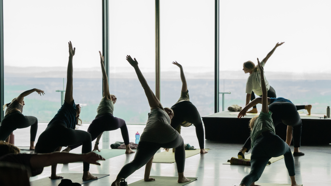 Neun Personen welche beim Yoga Brunch Yoga treiben. Eine Person ist auf einem Podest und zeigt die Übungen vor.