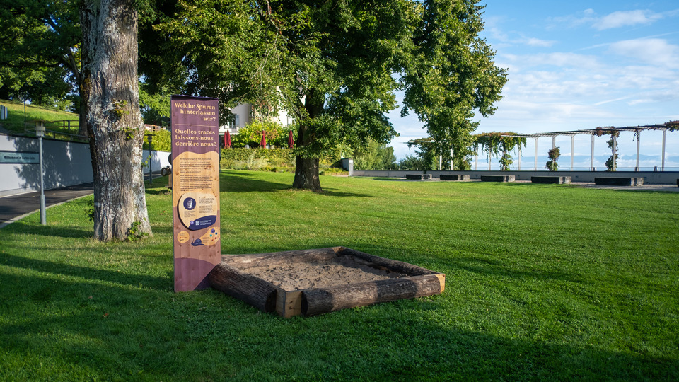Eine Stele vom Erlebnisweg «Natur. Und du?» mit einem Sandkasten und dem Kulm-Gebäude im Hintergrund.
