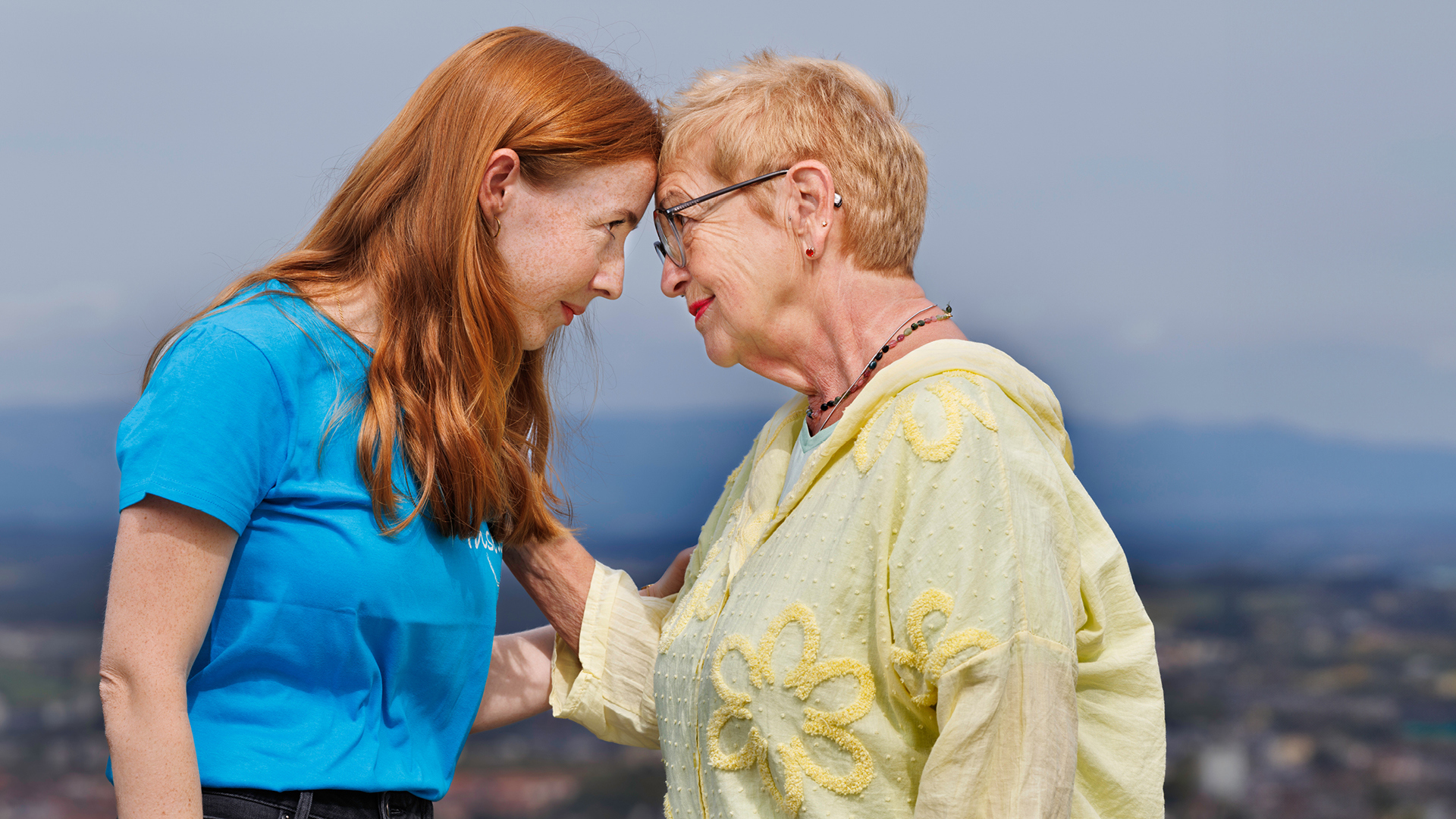 Eine junge rothaarige Frau und eine ältere Frau stehen Stirn an Stirn gegenüber voneinander. Dahinter die Aussicht vom Gurten.