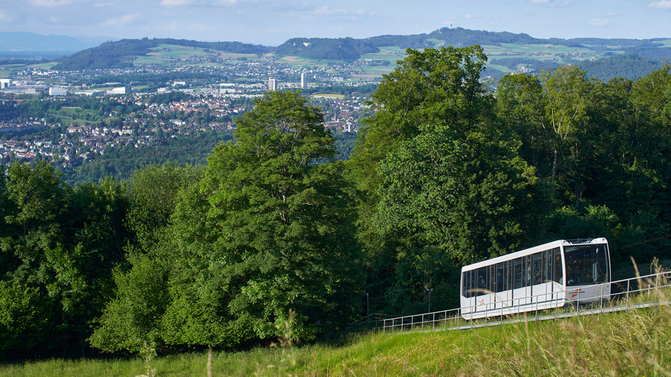 Eine Aufnahme der Gurtenbahn von oben, bei Tag, vor Bäumen mit Sicht auf die Stadt Köniz.