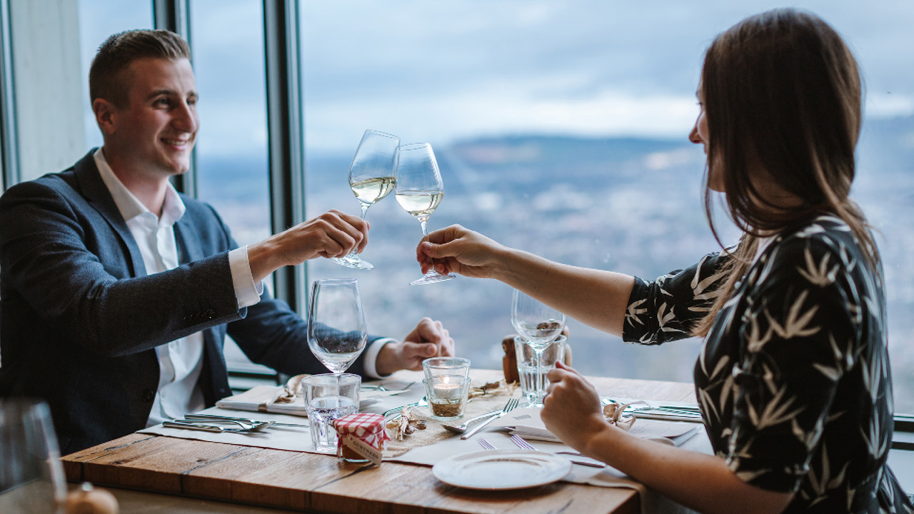 Zwei Personen welche an einem Tisch sitzen und beim Candlelight Dinner mit einem Glas Weisswein anstossen.