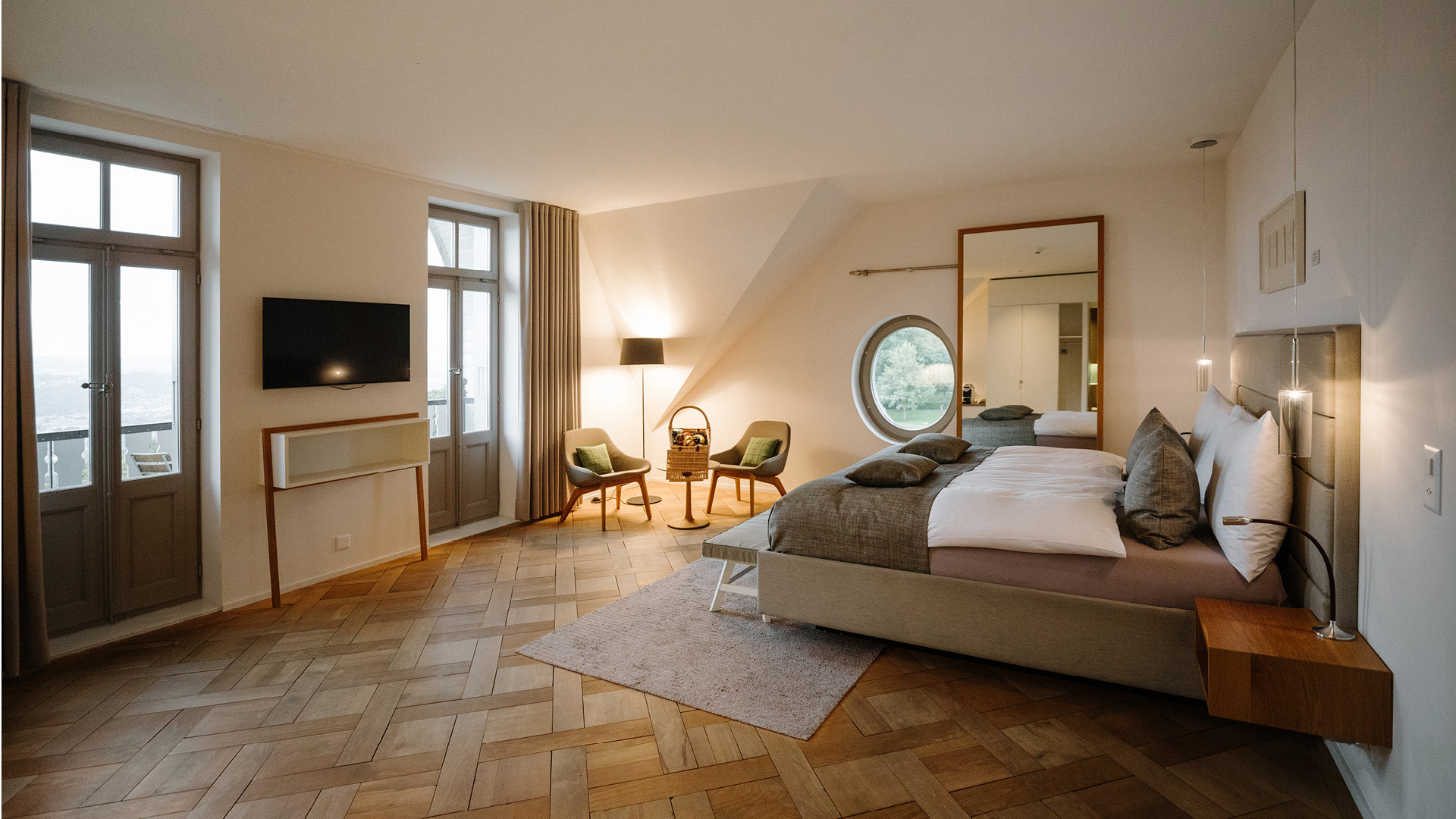 Ein Hotelzimmer mit zwei Balkontüren, einem grauen Bett, einem grossen Spiegel, einem runden Fenster und braunem Holzboden.