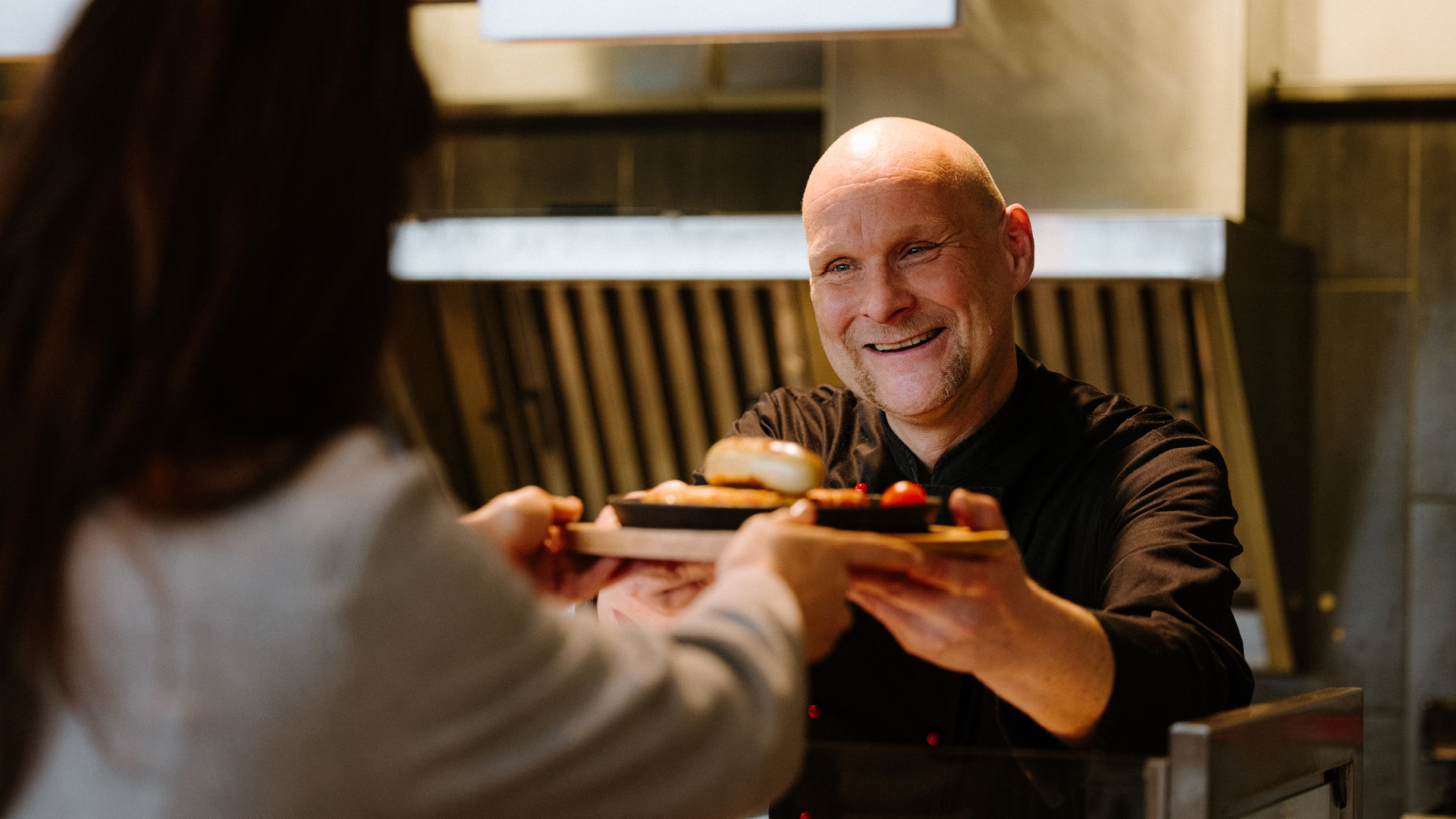 Ein Koch in einer schwarzen Kochjacke, welcher einem Gast gerade ein Teller über das Buffet in die Hand gibt.