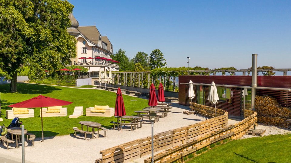 Das Restaurant Bahnhöfli. Es ist rot gestrichen, hat bei den Tischen rote Sonnensschirme und liegt neben einer grünen Wiese.